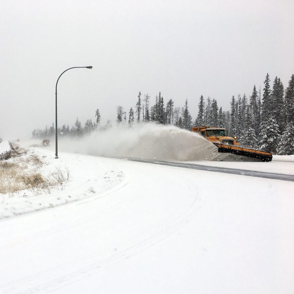 A Dawson Road Maintenance snowplow clearing Highway 97 in the Cariboo region of British Columbia, pushing a large cloud of snow off the roadway.