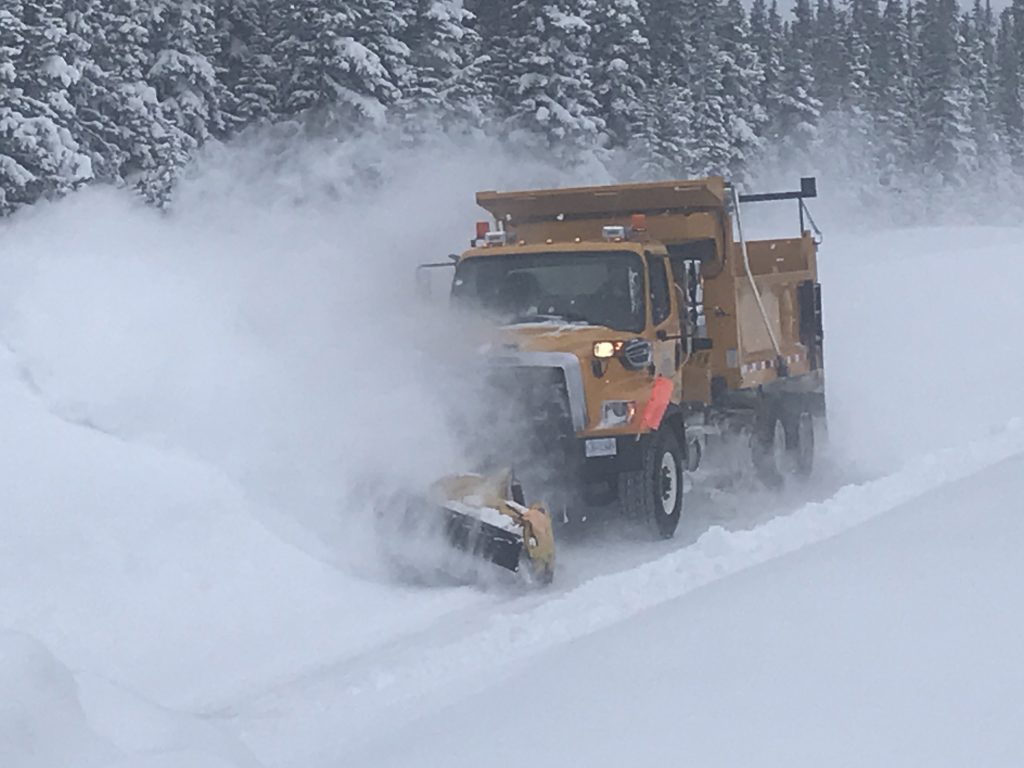 A Dawson Road Maintenance snowplow creating a snow cloud while clearing deep snow on a forested road in British Columbia.