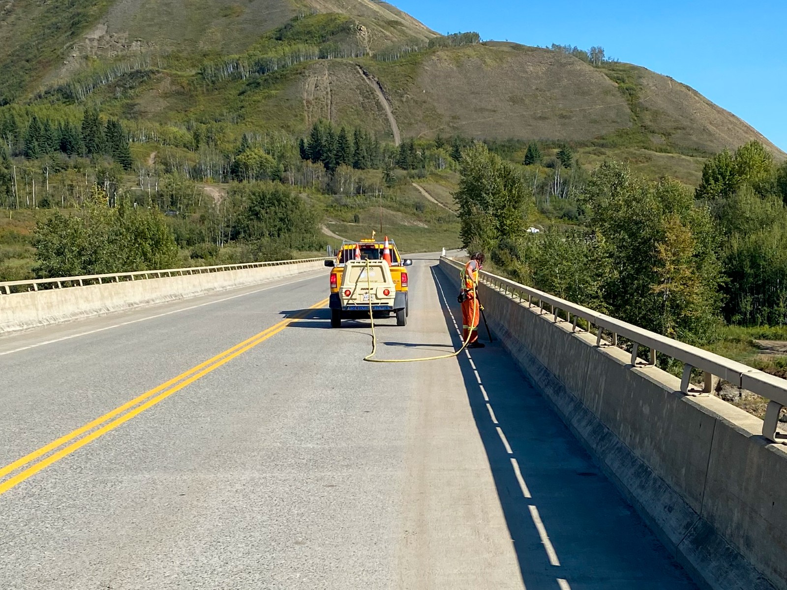 A worker in orange coveralls uses an air hose along the curb of a bridge deck, with a yellow work truck and trailer parked behind.